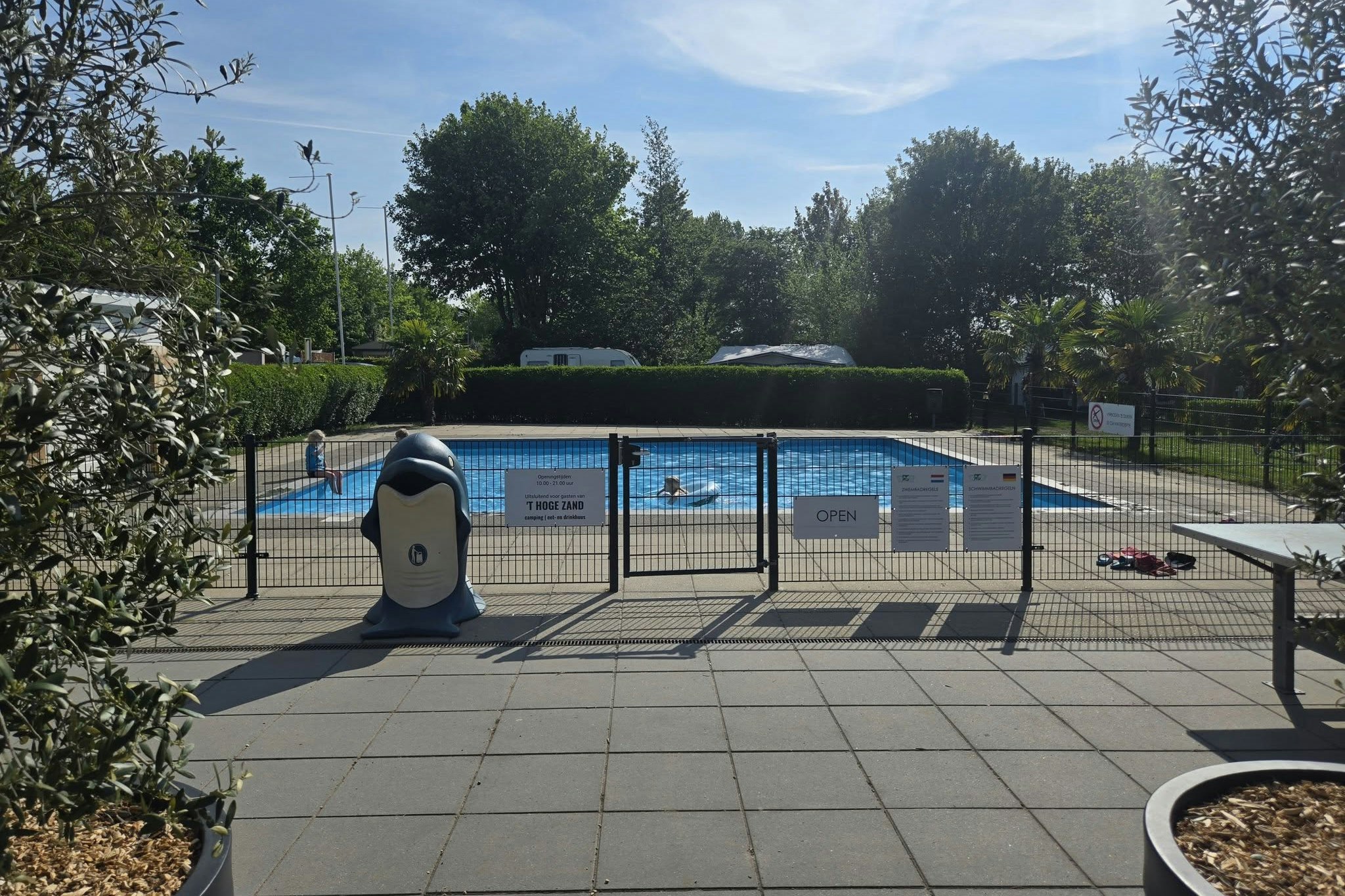 Beheizter Poolbereich mit umzäuntem Schwimmbecken, Sonnenliegen und gepflasterter Terrasse, umgeben von Bäumen und Sträuchern.