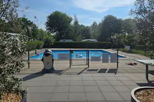 Beheizter Poolbereich mit umzäuntem Schwimmbecken, Sonnenliegen und gepflasterter Terrasse, umgeben von Bäumen und Sträuchern.