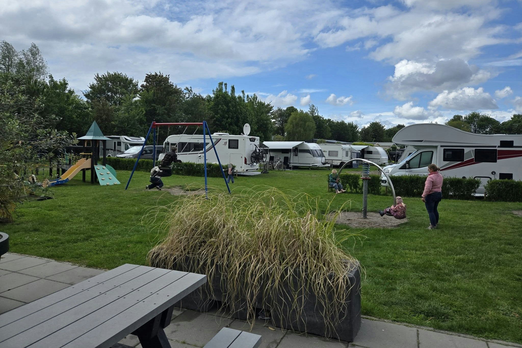 Campingplatz mit Wohnwagen und Zelten, Rasenfläche, Spielgerüst und Sitzbereich am Weg, im Hintergrund Bäume und Himmel.