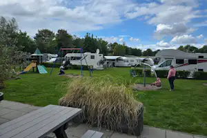 Campingplatz mit Wohnwagen und Zelten, Rasenfläche, Spielgerüst und Sitzbereich am Weg, im Hintergrund Bäume und Himmel.