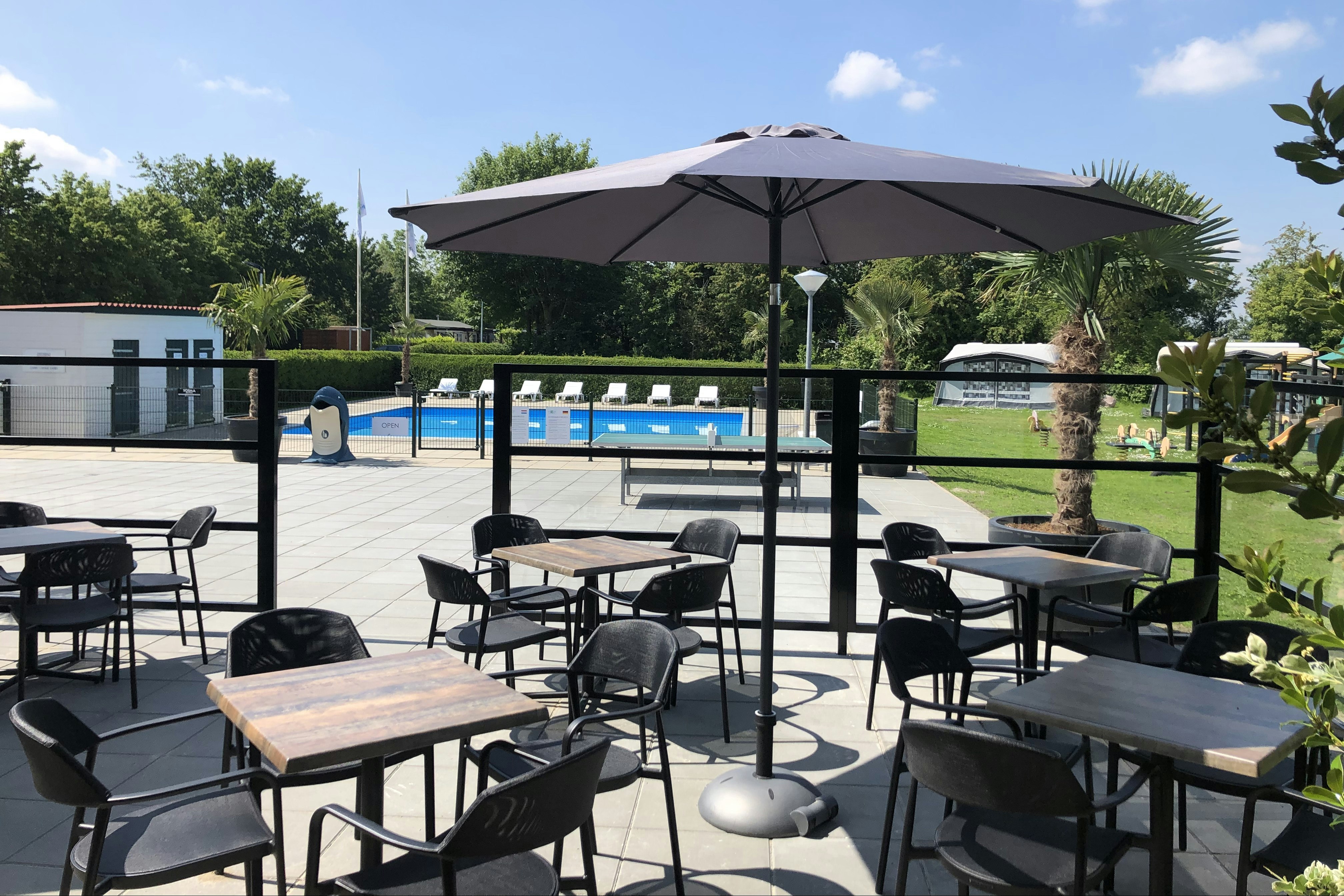 Terrasse mit mehreren schwarzen Gartenstühlen und Tischen, Sonnenschirm über dem Poolbereich, im Hintergrund Hecken und Gebäude.