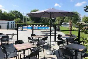 Terrasse mit mehreren schwarzen Gartenstühlen und Tischen, Sonnenschirm über dem Poolbereich, im Hintergrund Hecken und Gebäude.