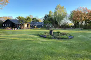 Grüne Campinganlage mit Rasenfläche, Blumenbeeten und mehreren Gebäuden im Hintergrund, umgeben von Bäumen und Wegen.