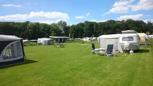 Wohnwagen und Zelt auf einem grünen Campingplatz, daneben Campingstühle und Tisch vor weiteren Stellplätzen im Hintergrund.