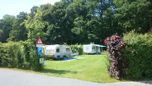 Wohnwagen auf einem Campingplatz-Stellplatz auf einer grünen Wiese, mit Hecke, Bäumen und Parkplatzschild am Weg.