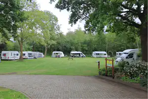 Stellplätze mit Wohnmobilen und Wohnwagen auf einer Rasenfläche, gepflasterter Weg und Bäume im Hintergrund.