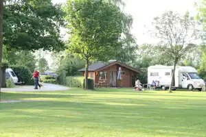 Holzhütte auf einem Campingplatz mit Rasenfläche, daneben Wohnmobil und weißem Transporter, Bäume und Weg im Hintergrund.