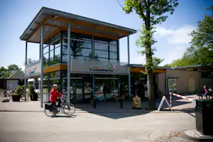 Camping-Rezeption mit Glasfront und Holzvordach, davor gepflasterter Platz, Fahrrad und Bäume vor dem Gebäude.