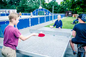 Tischtennis am Campingplatz: Kinder spielen auf einer blauen Tischtennisplatte, im Hintergrund Bäume und ein Gebäude.