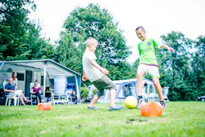 Zwei Kinder spielen mit einem Ball auf dem Rasen vor einem Wohnwagen und Campingstühlen im Grünen.