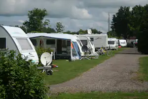 Reihe von Wohnwagen und Wohnmobilen auf einem Campingplatz, mit Grasstreifen, Kiesweg, Sitzgarnituren und Satellitenschüssel.