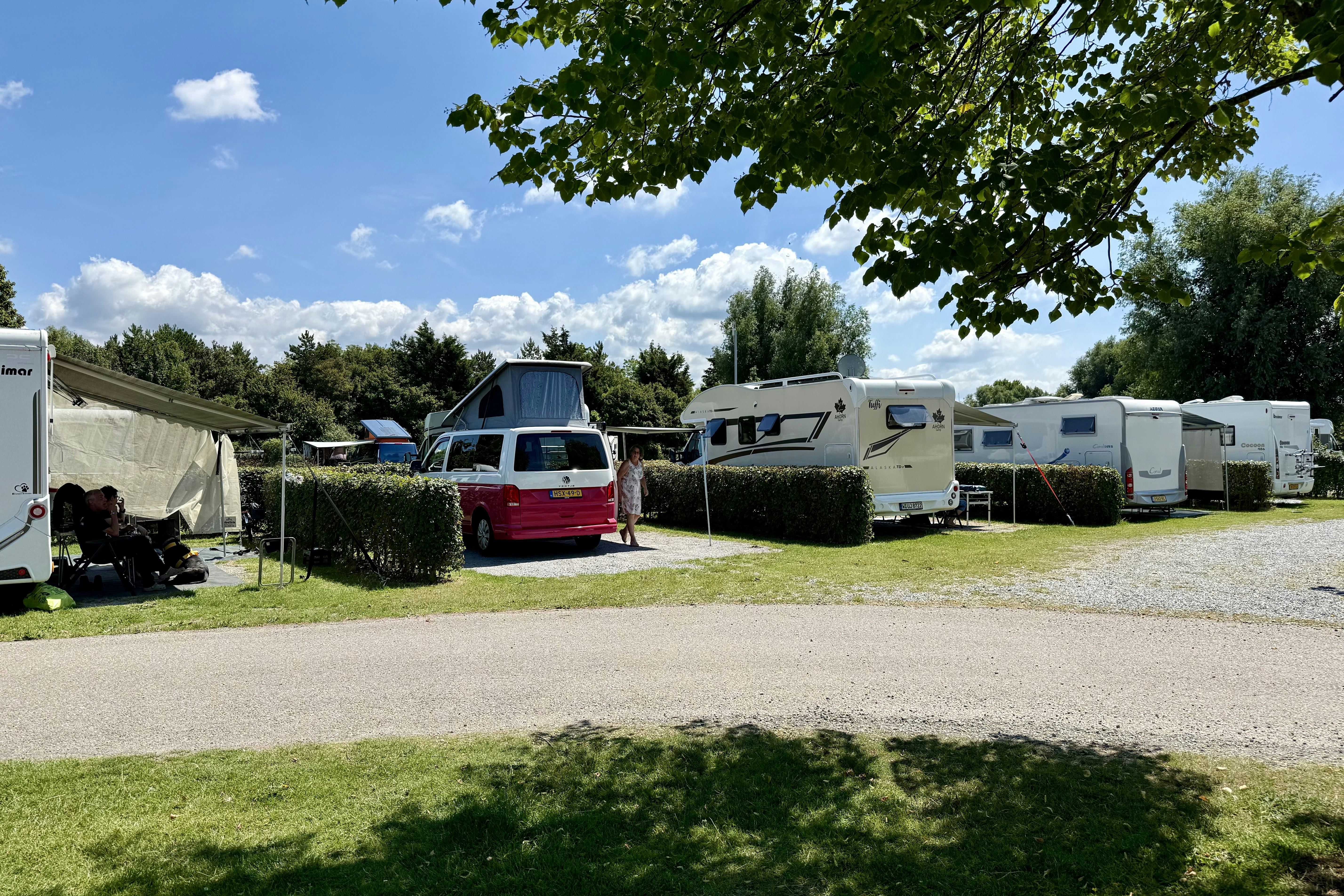 Wohnmobile und Caravans auf einem Campingplatz, mit Hecken, Rasenfläche und Kiesweg, im Hintergrund Bäume und Zelte.