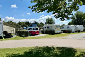 Wohnmobile und Caravans auf einem Campingplatz, mit Hecken, Rasenfläche und Kiesweg, im Hintergrund Bäume und Zelte.