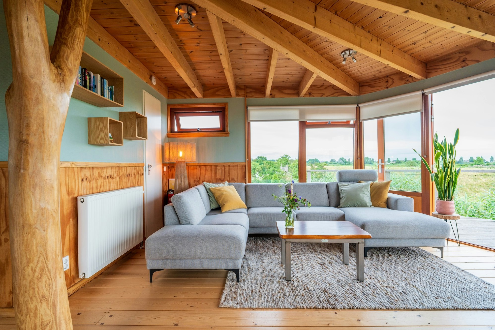 Gemütlicher Wohnbereich mit grauem Sofa, Couchtisch und Teppich unter Holzdecke, große Fensterfront mit Blick ins Grüne.