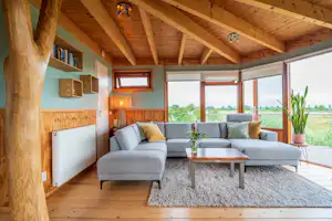 Gemütlicher Wohnbereich mit grauem Sofa, Couchtisch und Teppich unter Holzdecke, große Fensterfront mit Blick ins Grüne.