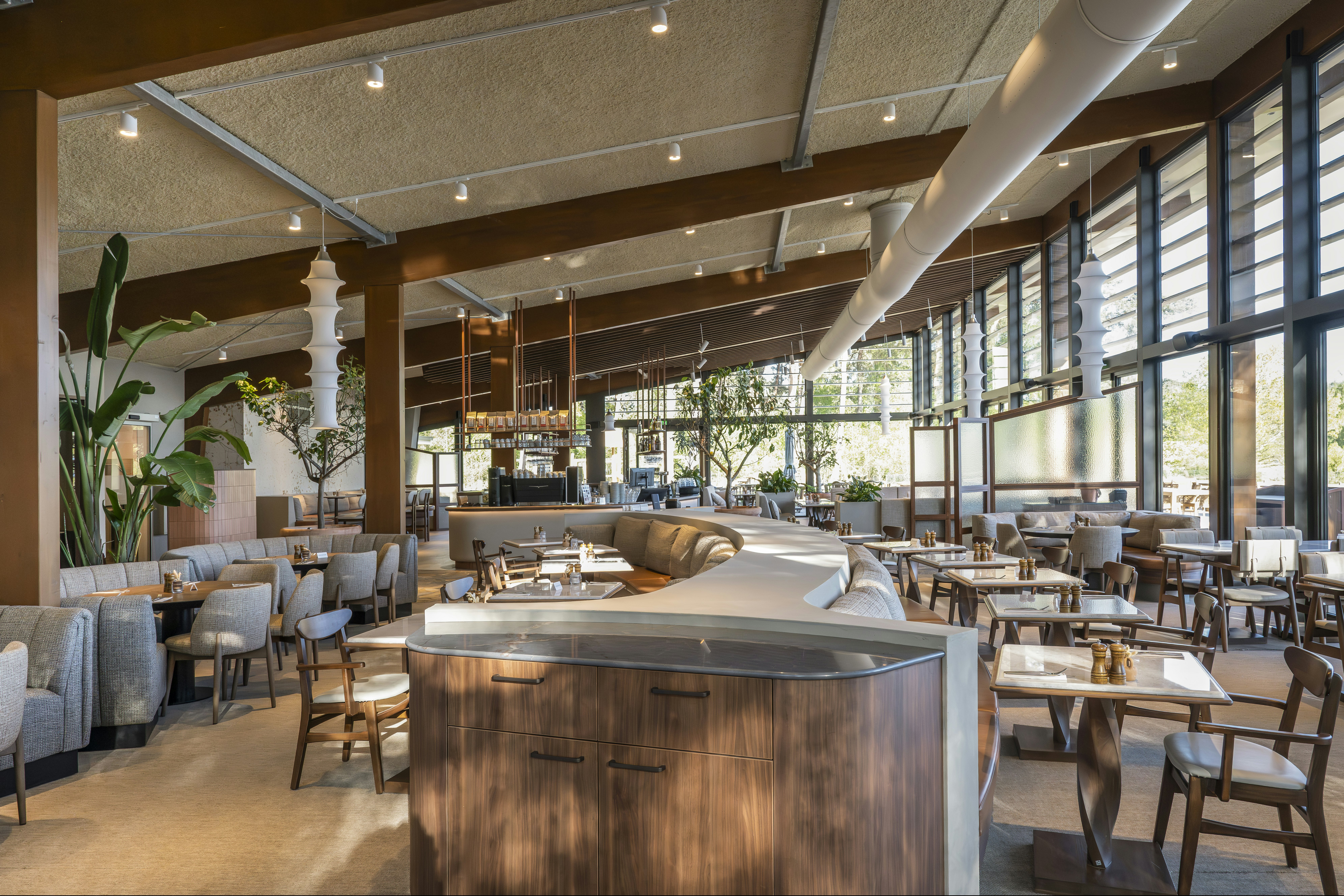 Innenbereich eines modernen Camping-Restaurants mit Theke, vielen Stühlen und Tischen, großen Fenstern und Pflanzen im Hintergrund.