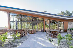 Große Terrasse mit Holztischen und Stühlen, Glasgeländer und Sonnenschirm, vor einem modernen Campinggebäude mit Bäumen im Hintergrund.