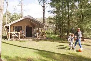 Zelt mit Holzpodest und Sitzbank am Campingplatz, umgeben von Bäumen und Rasenfläche, Kinder laufen im Vordergrund.