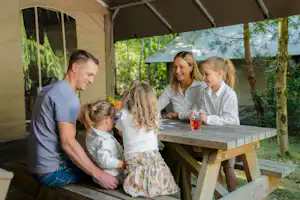 Familie sitzt an einem Holztisch unter einem Sonnenschirm im Campingbereich, im Hintergrund Bäume und Zeltwand.