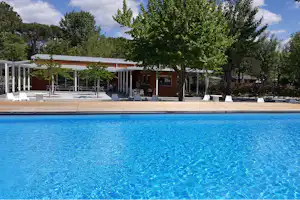 Blaues Camping-Schwimmbecken mit Holzterrasse, weißen Liegen und Bäumen vor einem Gebäude mit Veranda.