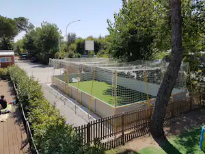 Kinder-Spielbereich mit Netzumrandung und grünem Boden, umgeben von Bäumen, gepflastertem Weg und Stellplätzen am Campingplatz.