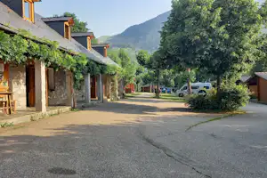 Campingplatz-Gasse mit Reihen von Holzgebäuden, Bäumen und einem Wohnmobil am Straßenrand, im Hintergrund Berge.
