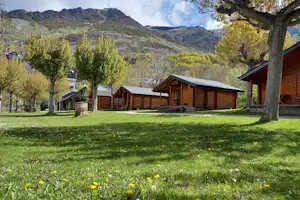 Holzhütten auf einem grünen Campingplatz, mit Bäumen, Rasenfläche und Blick auf die Berge im Hintergrund.
