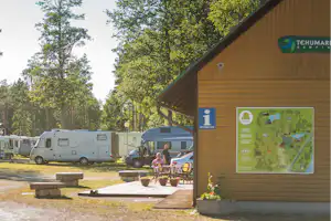 Wohnmobile auf dem Campingplatz neben einem Holzgebäude mit Infotafel und Sitzbereich, umgeben von Bäumen.