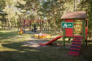 Spielplatz im Wald: Schaukelgestell, Rutsche und Sandkasten neben einem grünen Spielhaus auf dem Campinggelände.