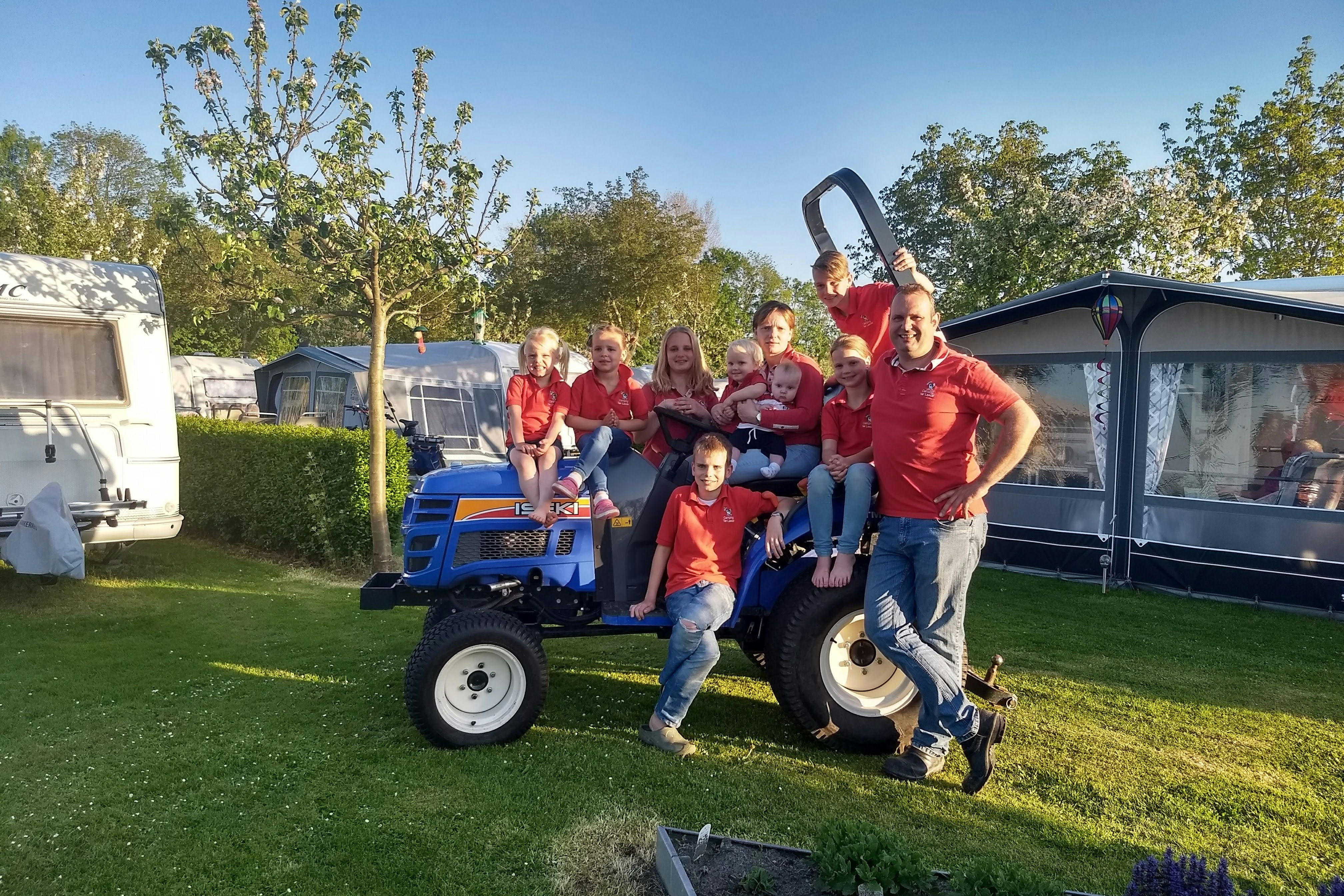 Familie auf dem Campingplatz vor einem Traktor auf dem Rasen, im Hintergrund Wohnwagen und Zeltanhänger.