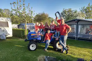 Familie auf dem Campingplatz vor einem Traktor auf dem Rasen, im Hintergrund Wohnwagen und Zeltanhänger.