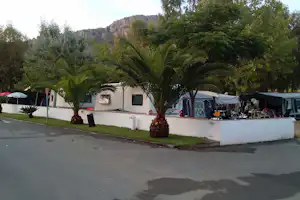 Wohnmobil-Stellplatz mit weißen Wohnmobilen, Palmen und Hecken vor einer Bergkulisse im Campingbereich.