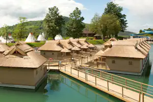 Campingdorf mit mehreren Strohdach-Hütten und einem grünen Pool, Holzsteg und Geländer am Wasser.