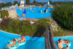 Buntes Außen-Schwimmbecken mit aufblasbaren Spielzeugen, Kinder und Erwachsene im Wasser, Steinmauer und Bäume im Hintergrund.