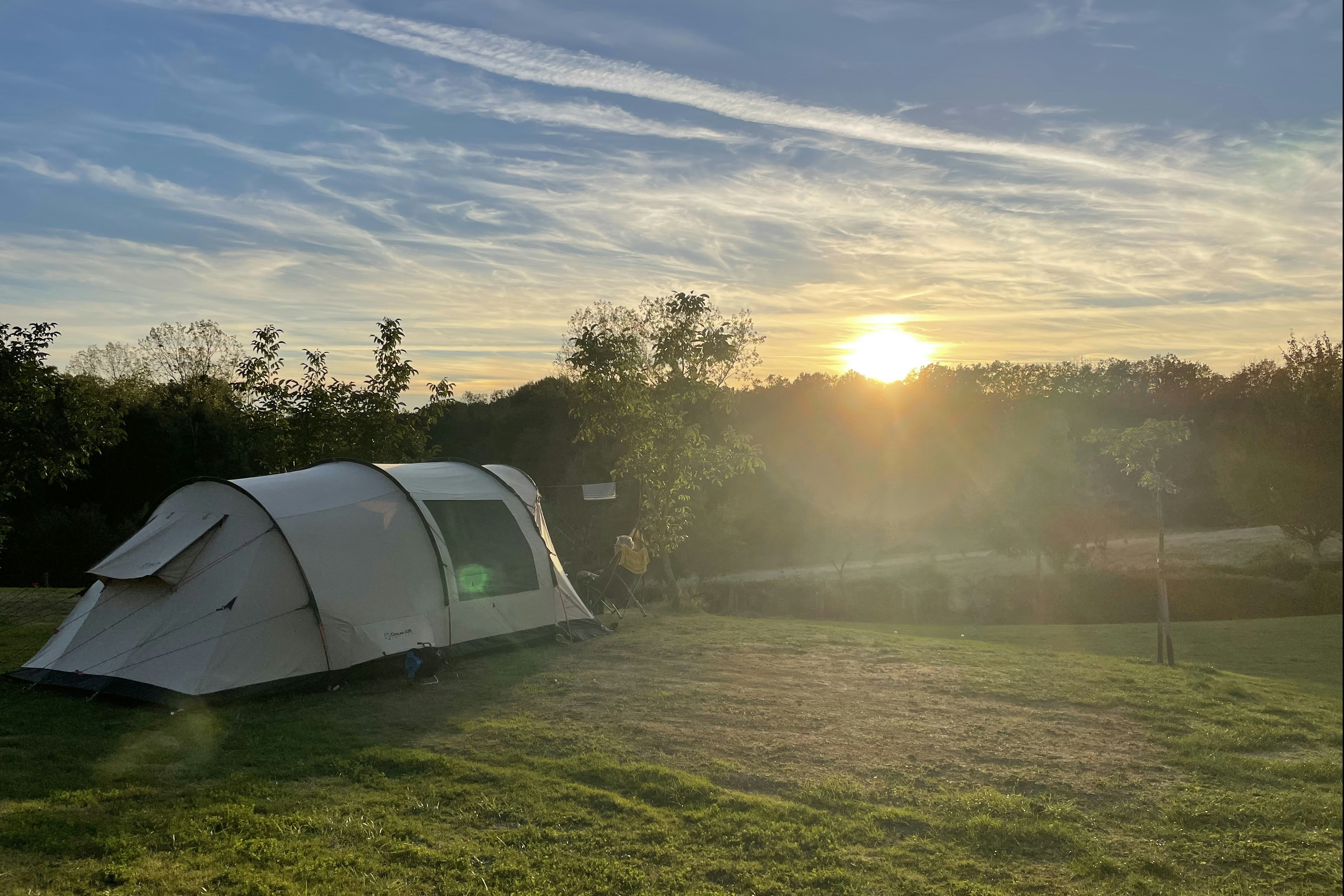 Zelt auf einem Campingplatz auf einer Wiese, im Hintergrund Bäume und die aufgehende Sonne am Himmel.