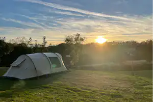 Zelt auf einem Campingplatz auf einer Wiese, im Hintergrund Bäume und die aufgehende Sonne am Himmel.