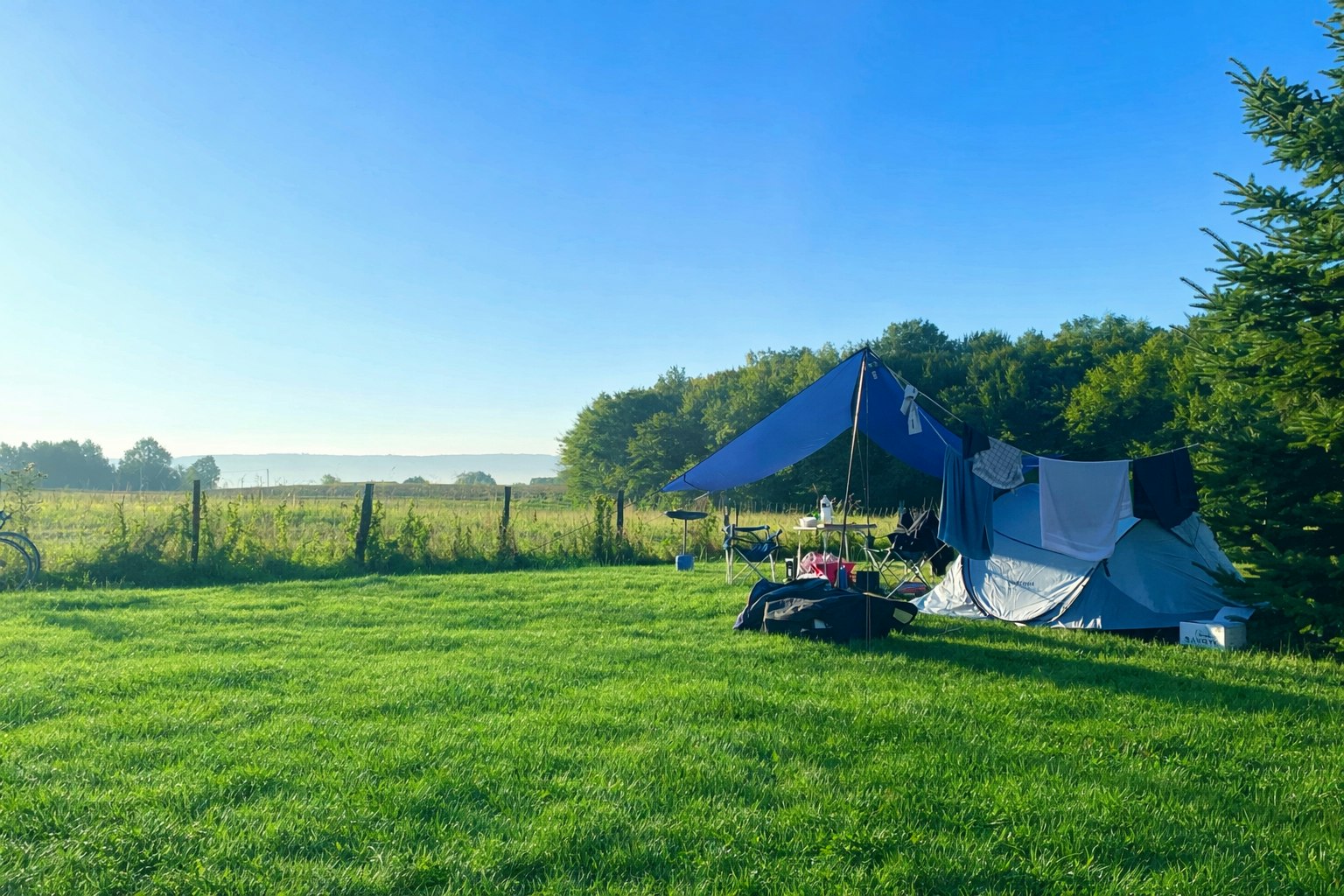 Campingzelt auf einer grünen Wiese am Waldrand, mit Blick auf Felder und einen klaren Himmel.
