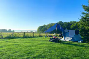 Campingzelt auf einer grünen Wiese am Waldrand, mit Blick auf Felder und einen klaren Himmel.