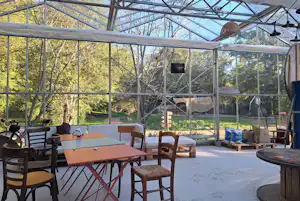 Überdachte Camping-Terrasse mit Holztischen und Stühlen, Blick durch Glaswände in den Wald und auf einen Gartenbereich.