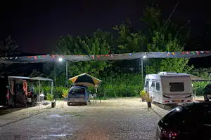 Campingplatz bei Nacht: Wohnwagen und Zelt unter Lichterkette, Kiesstellplatz, Bäume im Hintergrund, geparkte Autos.