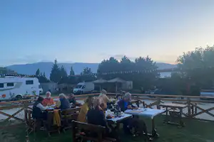 Camping-Terrasse mit mehreren Personen am Tisch, Holzzaun und Blick auf Berge und Bäume bei Abendlicht.