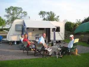 Wohnwagen mit Vorzelt und Terrasse, Campingstühle und Tisch auf Kiesplatz, daneben grüner Rasen und weitere Fahrzeuge.