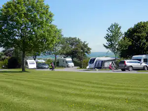 Campingwiese mit Wohnwagen und Zelt, daneben geparkte Autos und Bäume, im Hintergrund ein See.