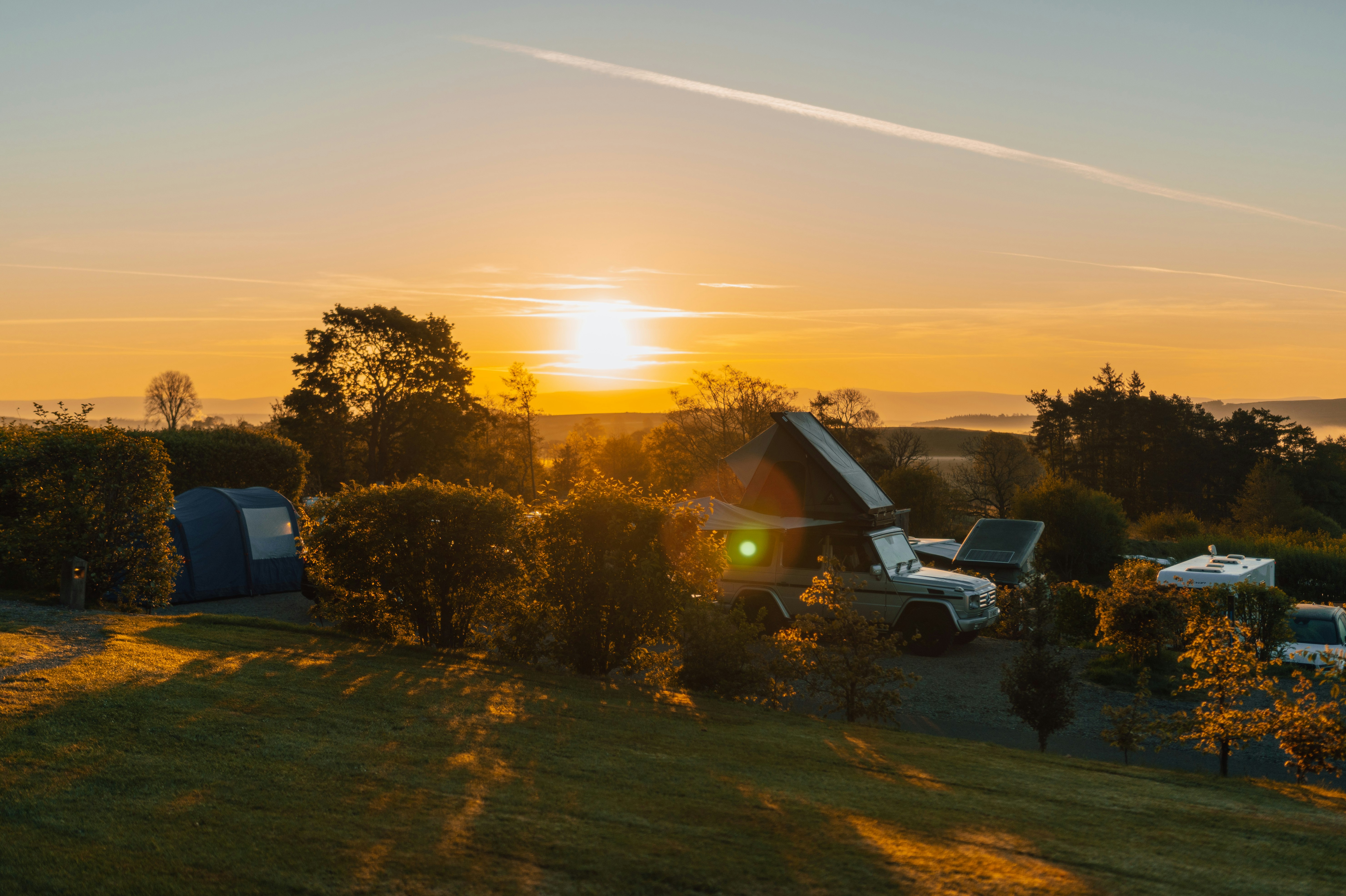 Campingzelt und Wohnmobil auf einer Wiese bei Sonnenuntergang, umgeben von Bäumen und Hügeln im Hintergrund.