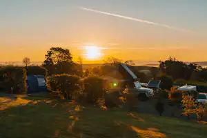 Campingzelt und Wohnmobil auf einer Wiese bei Sonnenuntergang, umgeben von Bäumen und Hügeln im Hintergrund.