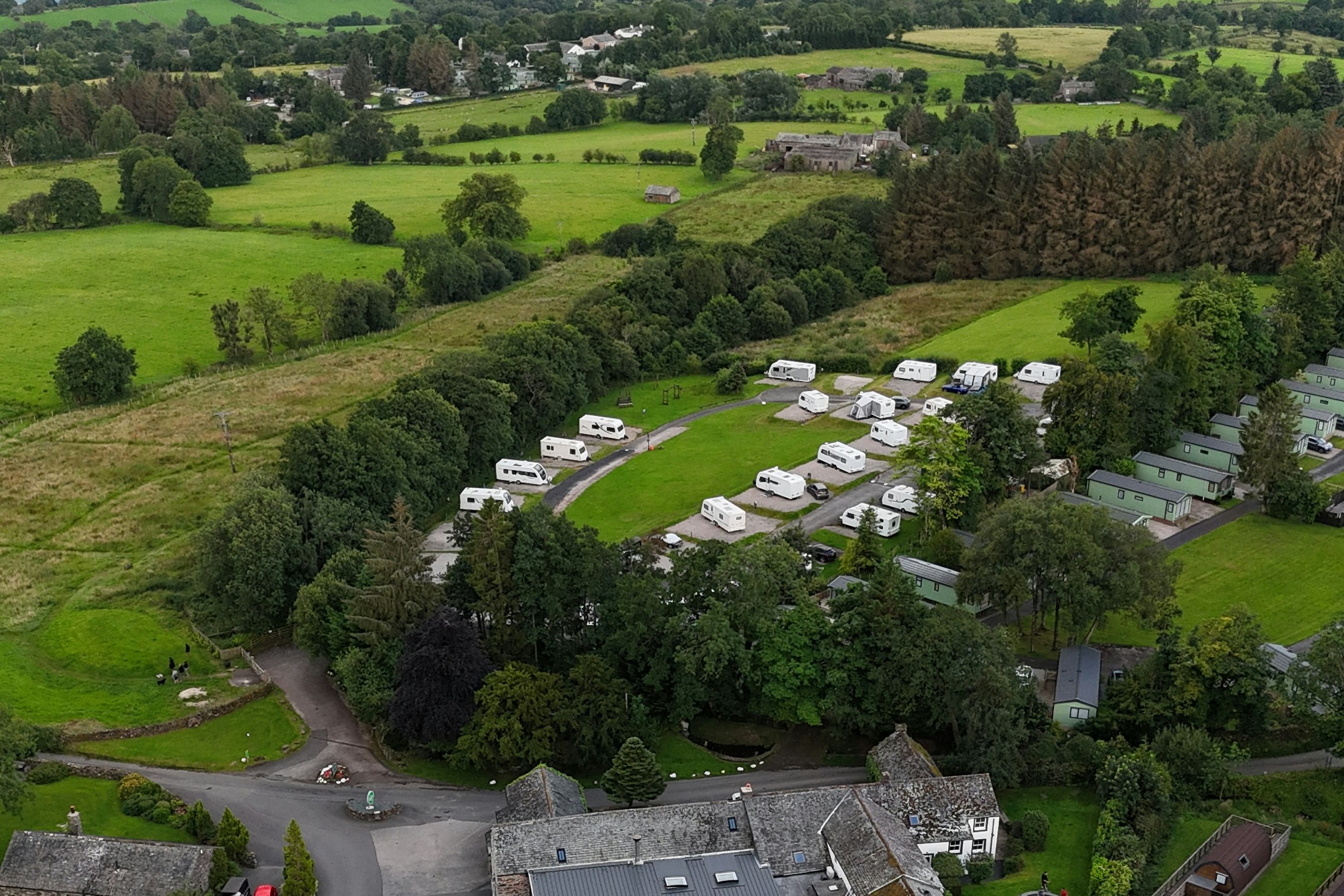 Luftaufnahme eines Campingplatzes mit Wohnwagen und Zelten in grüner Landschaft, Zufahrtsstraßen, Bäumen und einem See im Hintergrund.