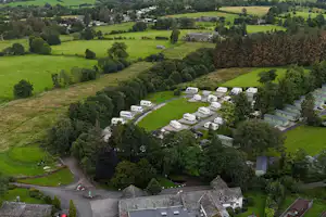 Luftaufnahme eines Campingplatzes mit Wohnwagen und Zelten in grüner Landschaft, Zufahrtsstraßen, Bäumen und einem See im Hintergrund.