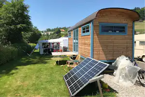 Holz-Mobilheim mit großen Fenstern, Terrasse und Solarmodulen auf dem Stellplatz, daneben Zelt und Wohnwagen.