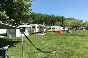 Campingplatz mit Wohnwagen und Zelten auf grünem Rasen, Markise, Campingstühlen und Bäumen im Hintergrund.