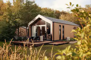 Modernes Holz-Campinghaus mit Terrasse, Sitzgruppe und Tisch im Grünen, umgeben von Bäumen und Sträuchern.